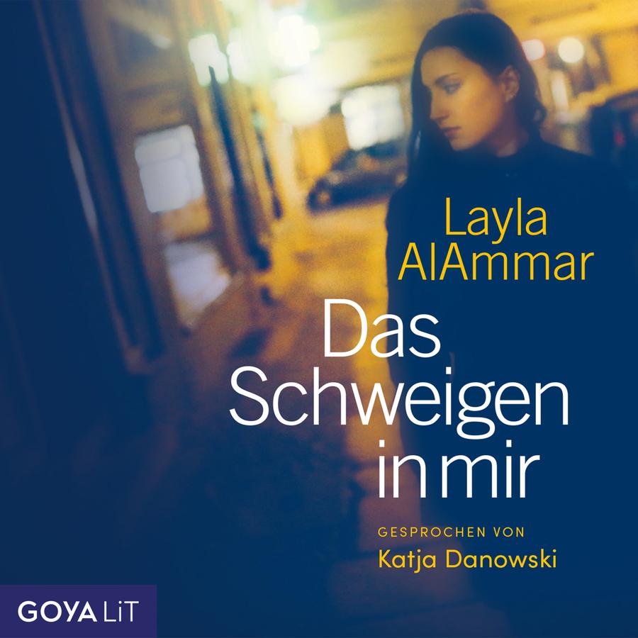 Das Schweigen in mir“ von Layla AlAmmar: Atmosphärisches Cover mit unscharfer nächtlicher Straßenszene in Blau- und Gelbtönen. Rechts steht eine dunkelhaarige Frau in dunkler Kleidung und blickt nach unten zur Seite. Autorinnenname und Titel stehen in gelber und weißer Schrift im rechten Bildbereich. Unten rechts steht „Gesprochen von Katja Danowski“, unten links das Logo „GOYA LiT“.
